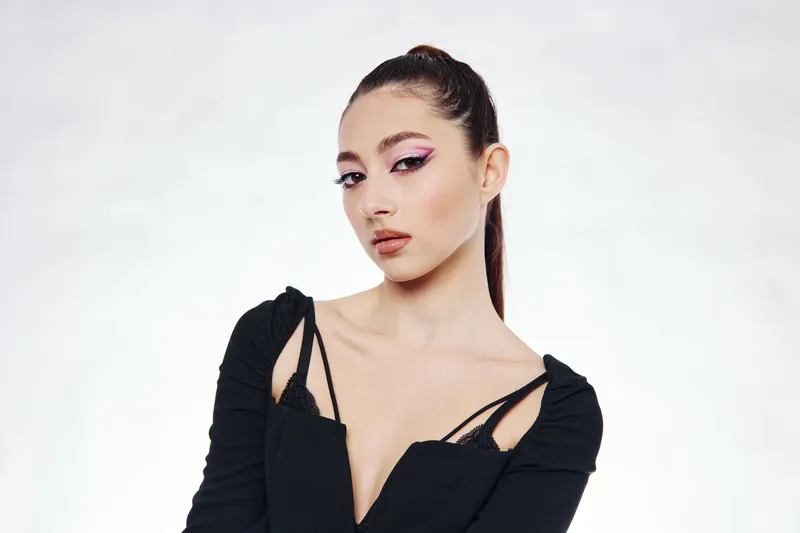 Young woman in creative makeup and black outfit aesthetically poses in front of a white background. Her confident gaze emphasizes fashion and beauty concepts with an edgy style.