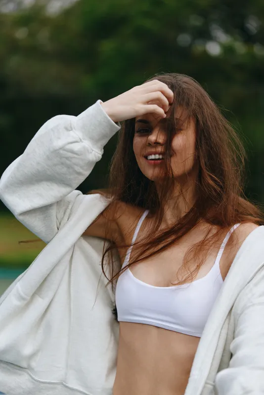 Young woman smiling and posing outdoors in a casual white outfit, showcasing natural beauty, confidence, and a carefree attitude in a lush green setting