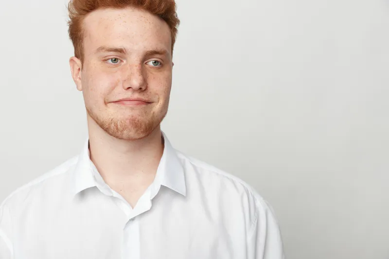 Young redhead man in white shirt looks sideways with skeptical expression on light background. Concept of doubt, uncertainty, and casual portrait of male model with freckles.