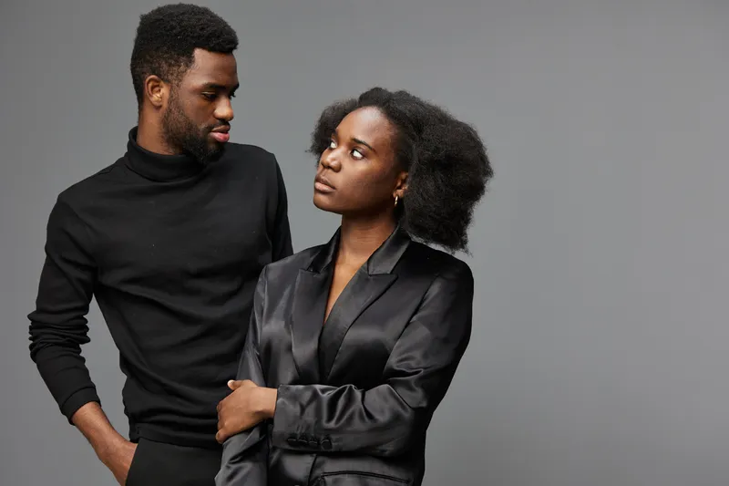 Man in black turtleneck and woman in satin blouse express deep emotions against a neutral gray background, showcasing modern fashion and intimacy