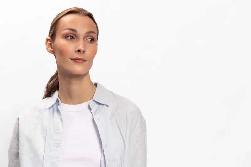 Young woman with light skin and brown hair looks to side wearing white shirt and light jacket. Studio portrait on white background. Concept for beauty, fashion, and lifestyle.