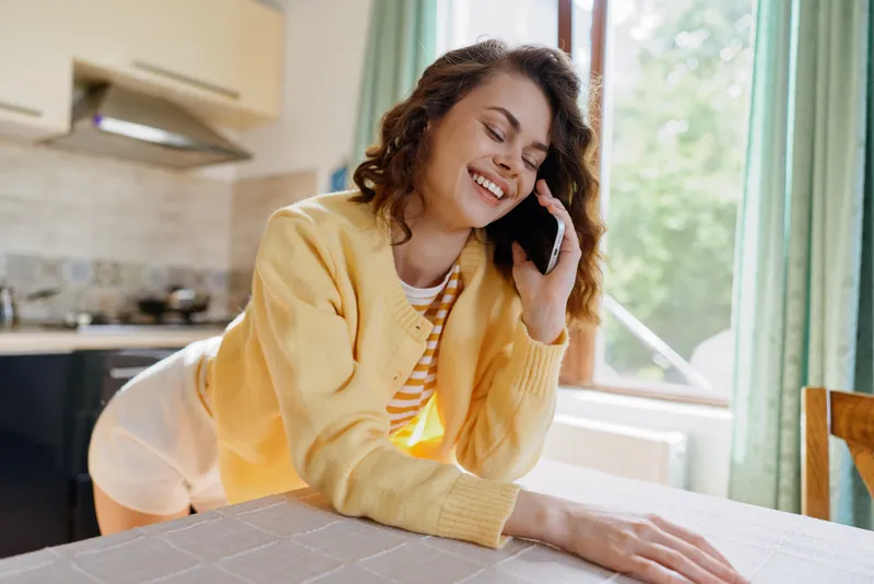 Young adult woman in a yellow striped cardigan chats on a smartphone at home, leaning over a table with a warm genuine smile beside a bright window and cozy kitchen.