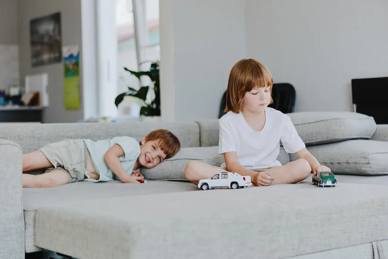 Two playful children on a cozy sofa enjoying their toy cars, one boy lying down and the other focused on playtime, radiating joy and innocence.