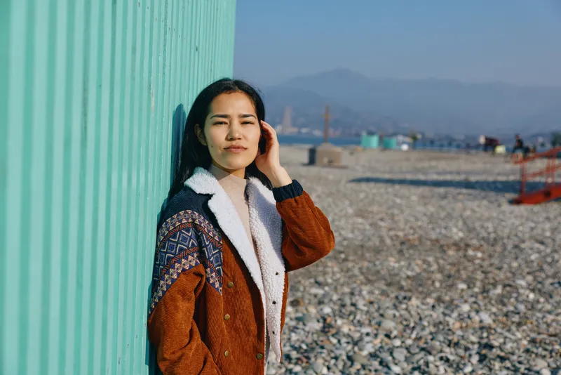 Woman in a knitted cardigan leans against a turquoise wall at a pebble coastal beach, distant city skyline visible, conveying a calm outdoor fashion moment with casual style and sunlit atmosphere