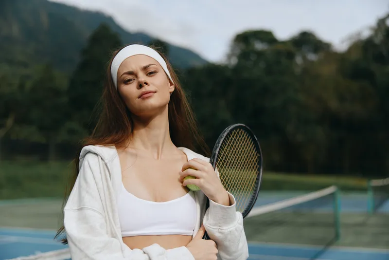 Young woman in athletic wear with a tennis racket, exuding confidence and determination on a tennis court surrounded by lush greenery and soft natural lighting