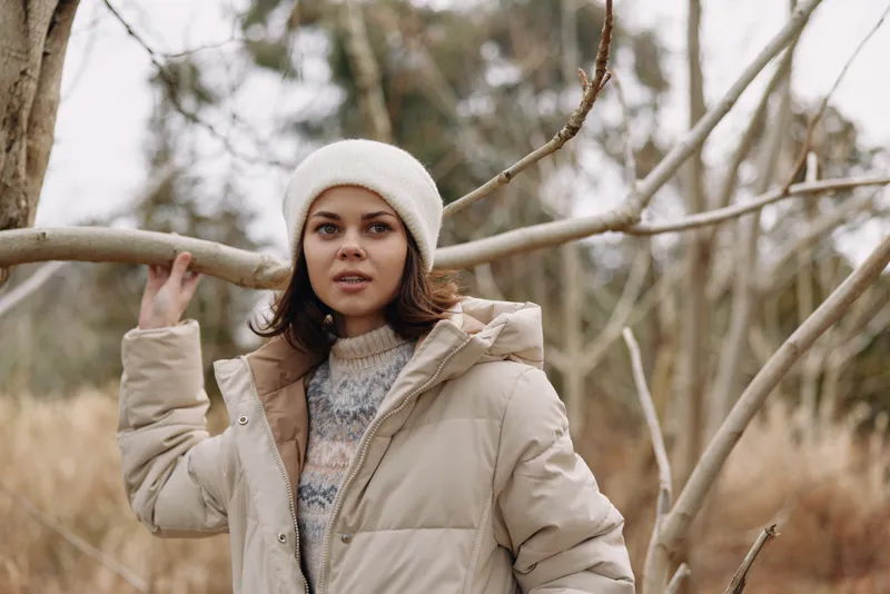 Winter scene with a thoughtful woman in a beige puffer jacket standing among bare branches, soft natural light, calm outdoors, cozy clothing and a serene mood.