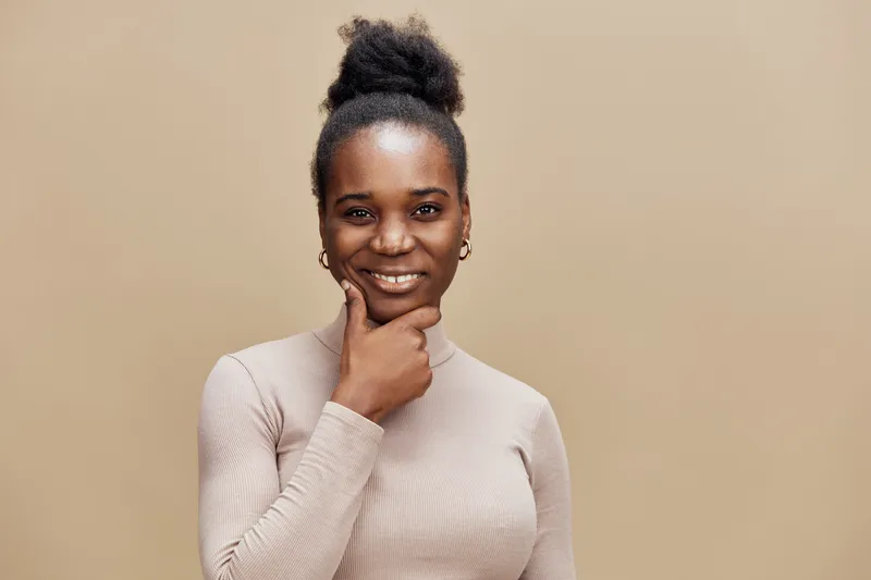 Smiling African woman in beige outfit looking confident and thoughtful, set against a soft brown background, ideal for concepts of empowerment and self care