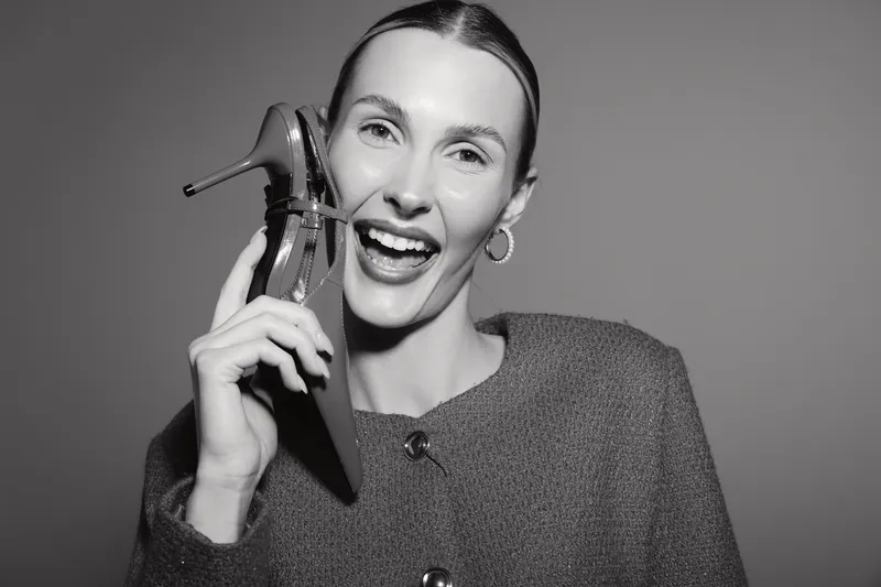 Happy Caucasian woman with fair skin holds shoe near face smiling joyfully. Studio portrait in black and white with playful and fashionable concept.