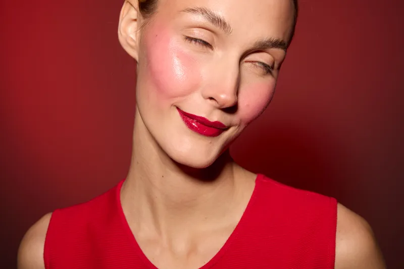 Close-up of a smiling woman with glossy red lipstick and bright pink blush on her cheeks wearing a red sleeveless top. Concept of makeup, beauty, fashion, and confidence.