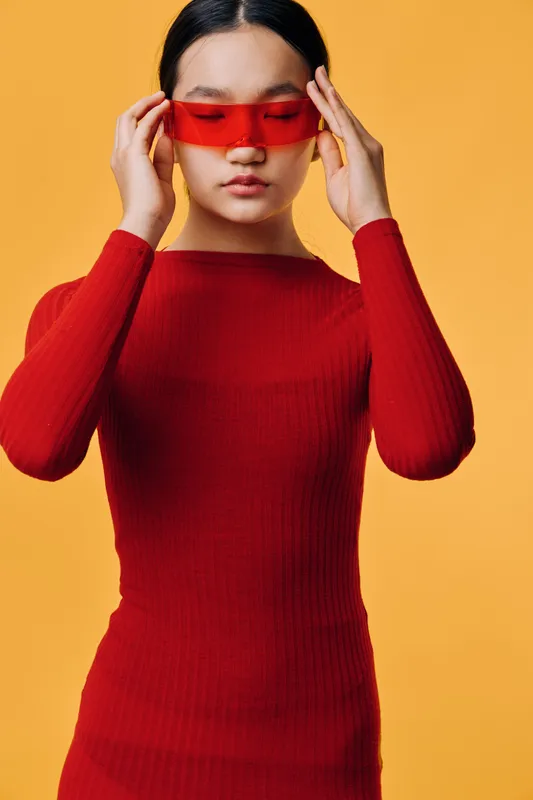 Woman in red sweater adjusting red translucent glasses on face with closed eyes against orange background. Fashion, portrait and style with casual clothing and modern accessory.