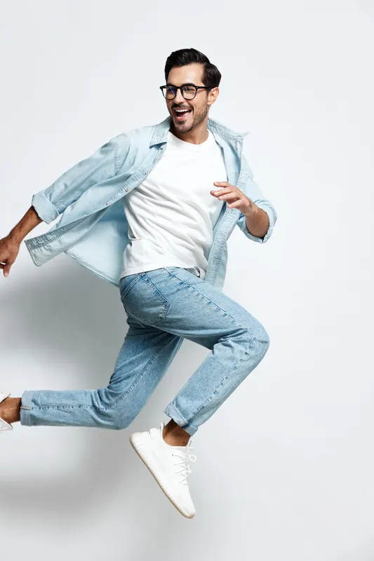 Energetic smiling man wearing denim jacket and glasses joyfully jumping against plain grey background, expressing happiness and vitality. People lifestyle concept.