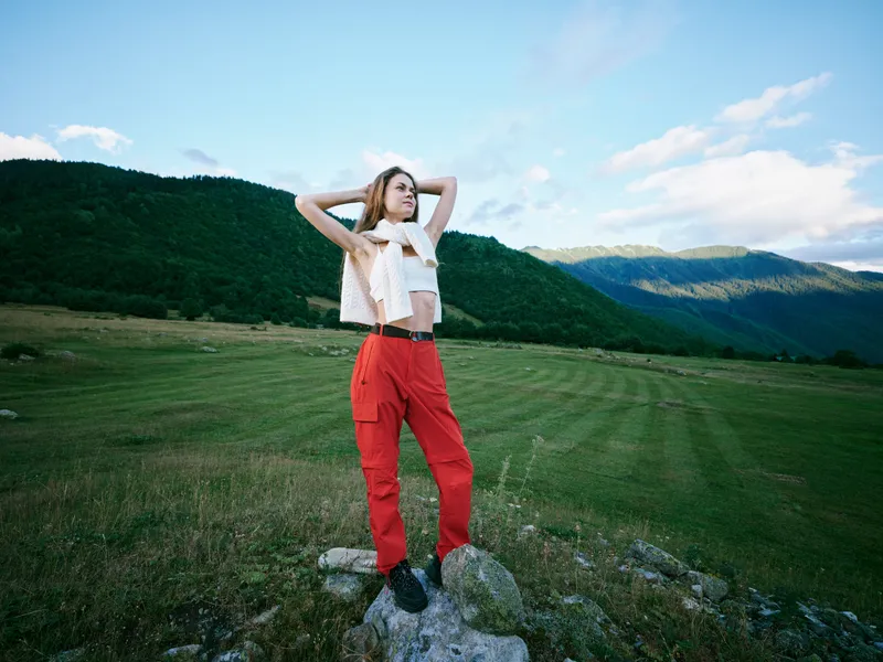 Outdoor field with mountains in the distance, a fashion pose on a rocky ledge, bright sunlight, red pants and white top, conveying freedom, adventure and a carefree summer mood in wide open space