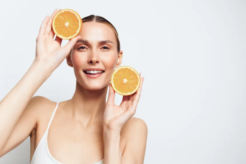 Smiling woman holding orange halves near face, fresh citrus concept, healthy skincare and wellness, bright light studio background, clean diet nutrition message for women