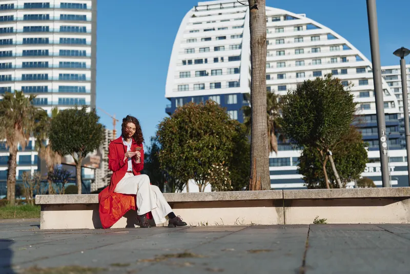 Young woman with curly hair sits on a concrete bench in a sunny urban park, wearing a red coat over a white dress, exuding calm confidence with modern buildings behind and a soft breeze.