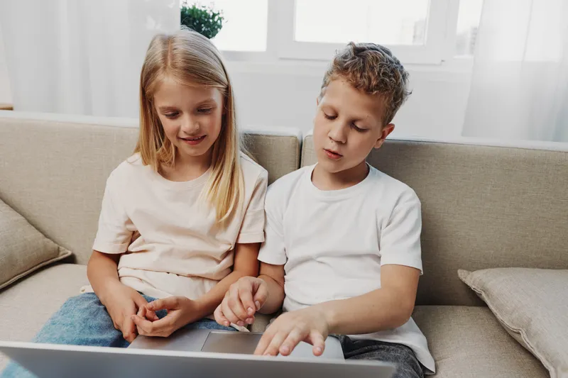 Two children sitting on a sofa using a laptop together, focused on the screen. Casual home environment with natural light from a window in the background.