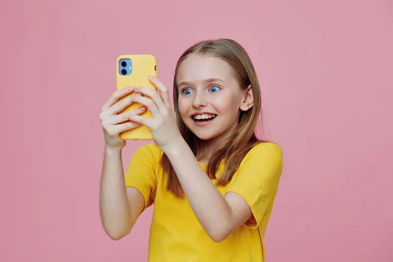 Happy young girl with blue eyes taking a selfie on a pink background, wearing a yellow shirt, expressing excitement and joy while using a smartphone