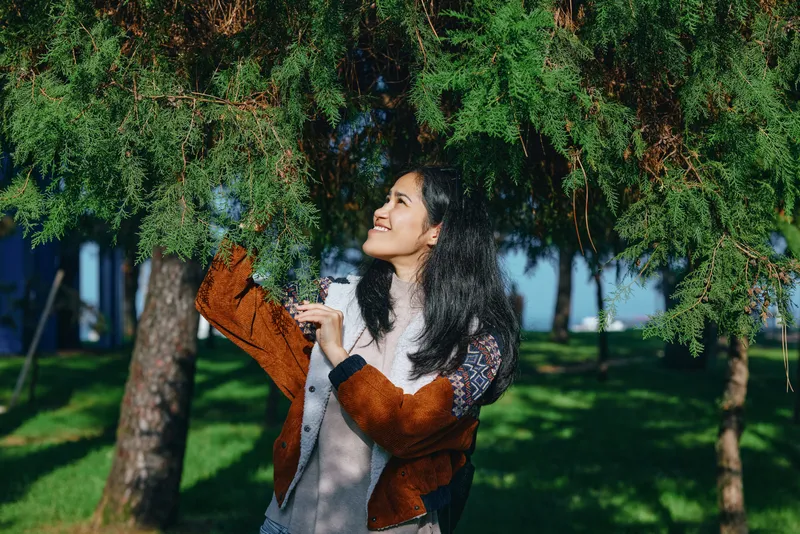 Woman sprays water under a lush tree in a sunny park, enjoying outdoor nature moment, casual clothing, cheerful expression, gardening activity, refreshing atmosphere