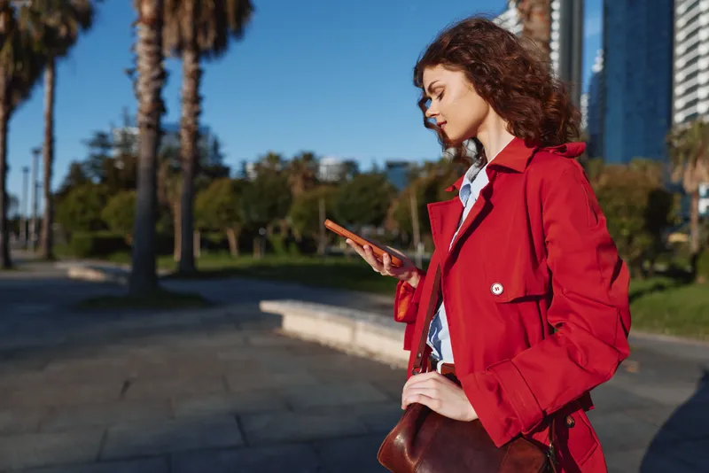 Young woman in a red coat stands outdoors on a sunny day, looking at a tablet in a modern urban park with a brown crossbody bag nearby