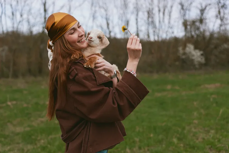 Woman dog outdoors flower smile happiness, casual portrait of a brown jacket wearer holding a small dog and a blossom in a sunny park, expressing warmth and affection in lifestyle photography