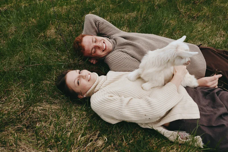 Two smiling people lie on grass, softly cradling a white goat kid. Casual sweaters, warm afternoon light, relaxed lifestyle. Connection with nature, friendship, and comfort in a peaceful meadow scene.