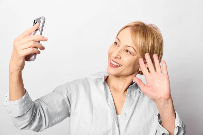 Cheerful woman taking a selfie with smartphone, smiling and waving hand, relaxed portrait in studio on white background, soft light, communication mood