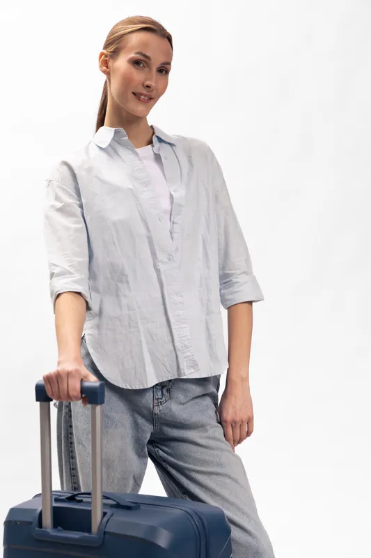 Young woman in casual clothes stands with blue suitcase on white background. Travel concept with confident female traveler ready for trip or vacation. Studio shot with soft lighting and natural pose.