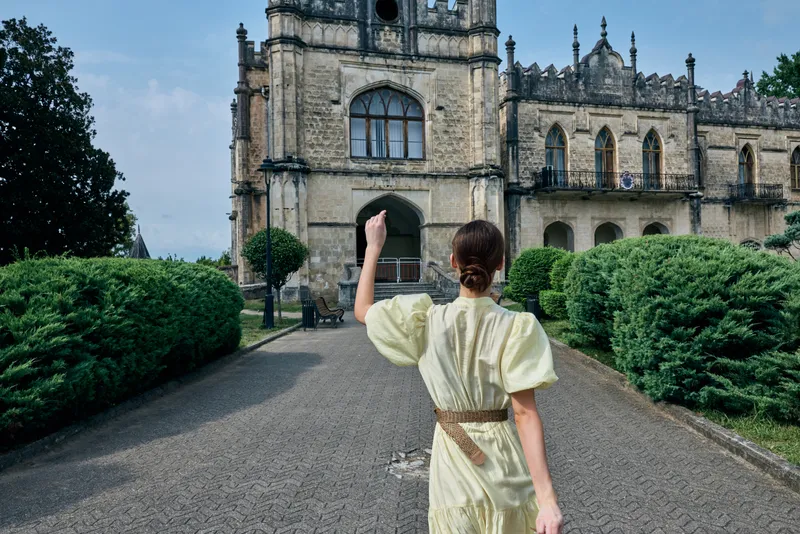 A woman in a pale yellow dress walks along a gravel pathway toward a historic castle, surrounded by manicured gardens, creating a calm travel moment outdoors