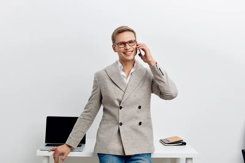 Confident professional man model standing in modern office with white wall background, smiling and talking on mobile phone, dressed in smart casual blazer and shirt, holding a tablet and working. Busi