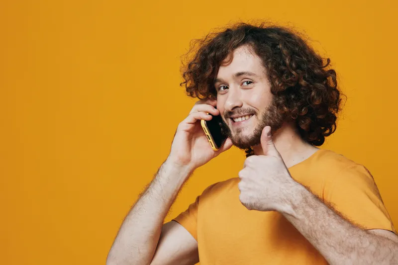 Happy young man with curly hair talking on smartphone against a bright yellow background, showing a thumbs up for success, conveying a positive vibe