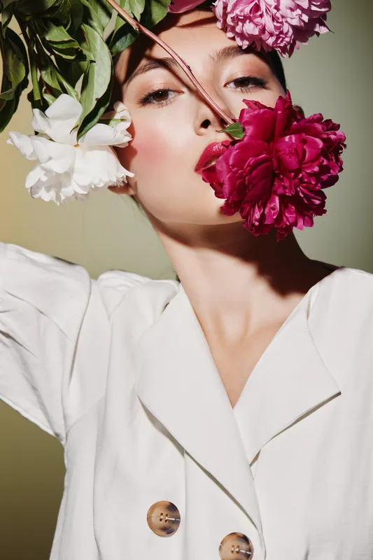 Fashion model with natural makeup holds large pink and white flowers close to face, wearing white blazer. Soft light emphasizes fresh beauty and floral creative styling concept.
