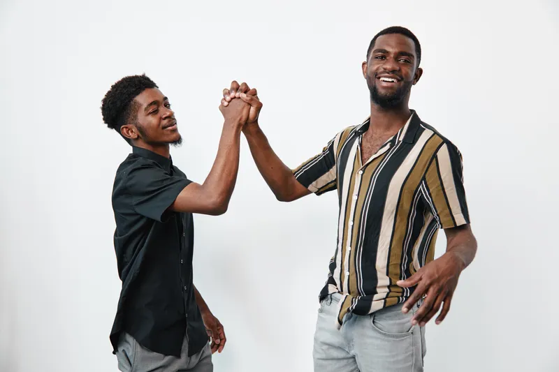 Two African American men with dark skin smiling and greeting each other with a handshake in casual clothes on white background. Friendship and connection concept.