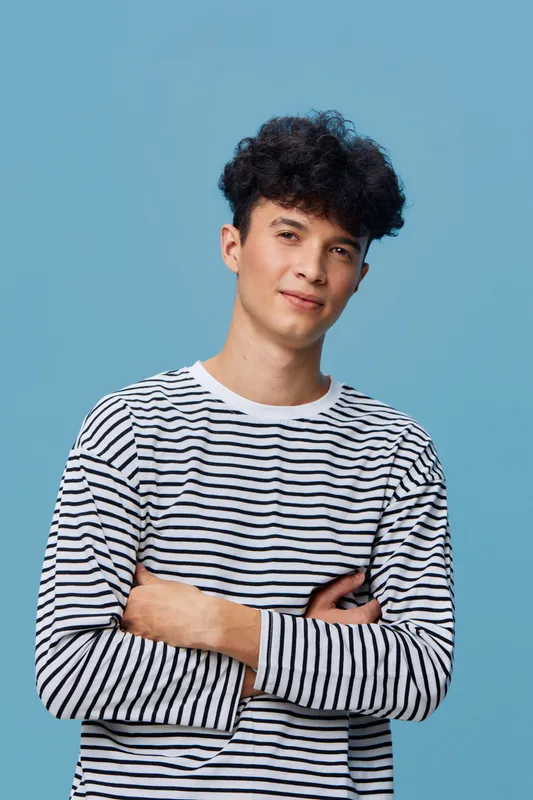 Young man with curly hair wearing a striped shirt stands confidently against a blue background, showcasing a casual and relaxed style with a calm expression.