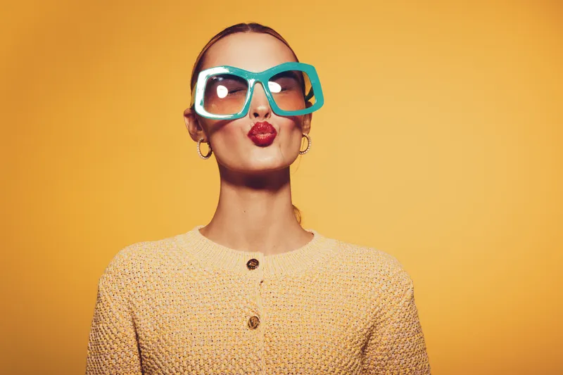 Stylish woman with oversized blue sunglasses and red lipstick makes a kiss face. She wears a beige knitted sweater against a bright yellow background. Concept of fashion and fun.