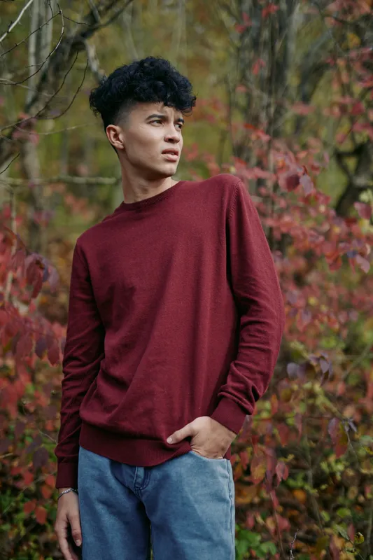 Young man in a burgundy sweater stands amid autumn foliage, natural candid pose conveying authenticity and credibility in a portrait, subtle expression and relaxed body language.