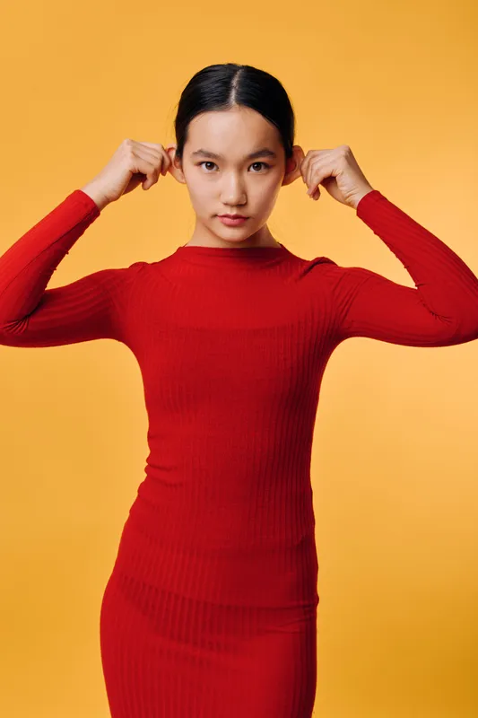 Young woman wearing red dress posing with hands touching hair against orange background, confident expression with straight posture, studio portrait with vibrant colors.