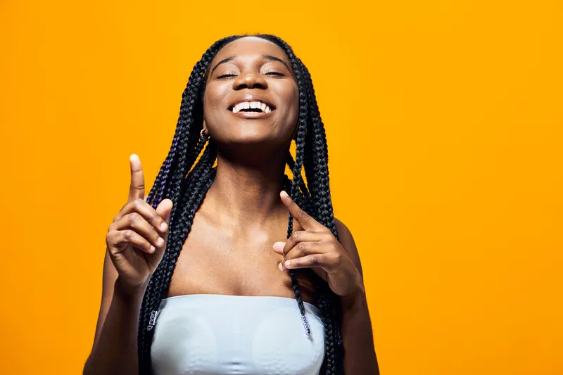 A joyful young African American woman with braided hair smiles brightly against a vibrant yellow background, expressing happiness and confidence in a stylish outfit