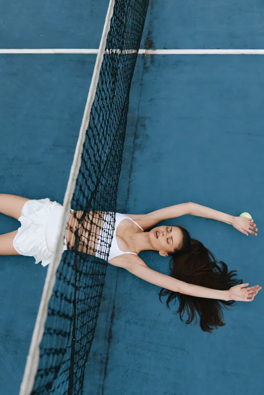 Young female tennis player relaxing on the court, dressed in a white sports outfit, with a cheerful expression, perfect for wellness and active lifestyle themes