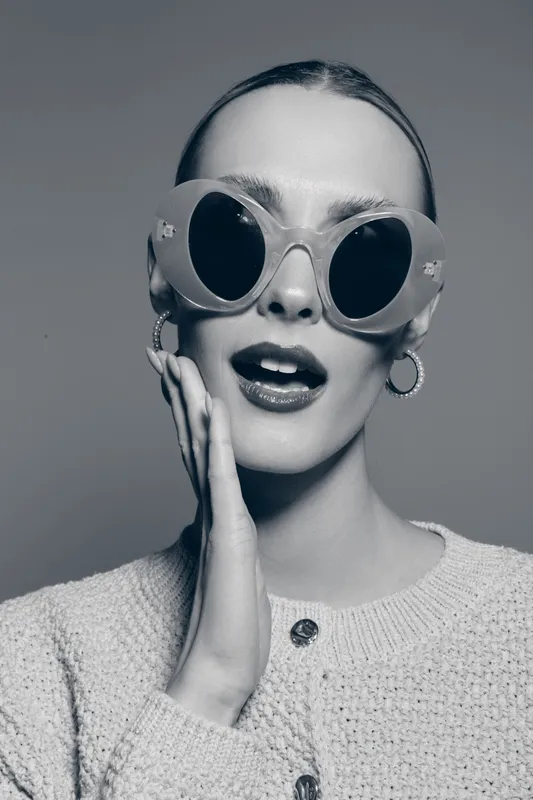 Elegant white woman with pale skin wears large round sunglasses and hoop earrings, touching her face with an open mouth. Concept for fashion, style, and beauty portrait in studio.