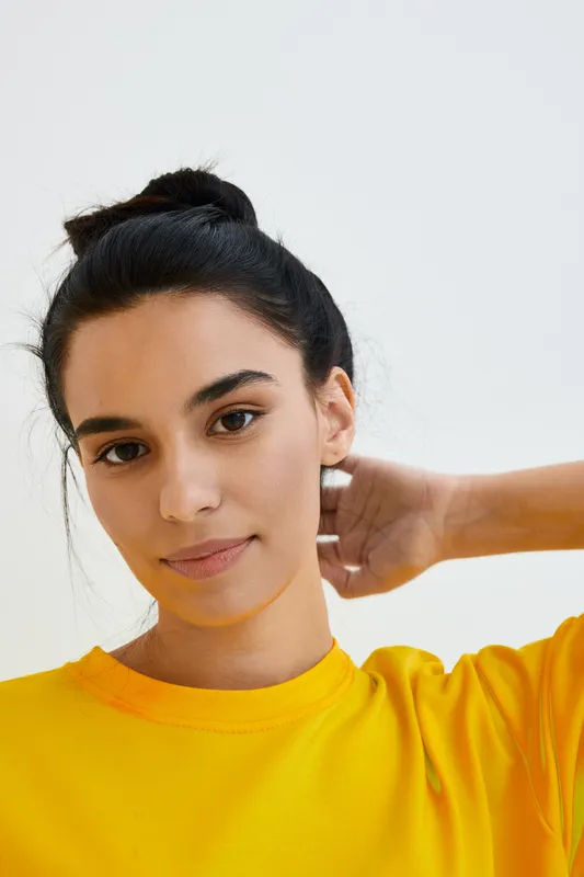 Young woman with dark hair in a yellow sweatshirt poses casually against a minimalist white background, showcasing natural beauty and relaxed confidence.