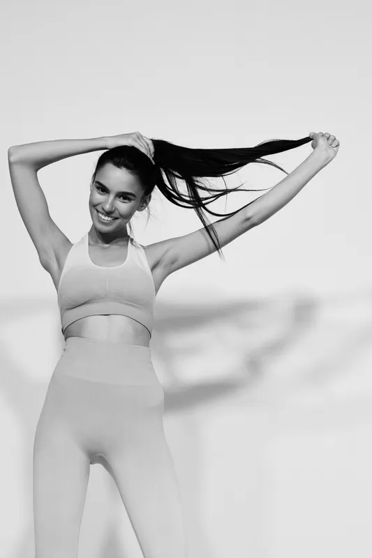 Sporty woman fitness smiling, holding her hair up, wearing athletic outfit, posing confidently in bright studio. Black and white fitness portrait with energetic vibe.