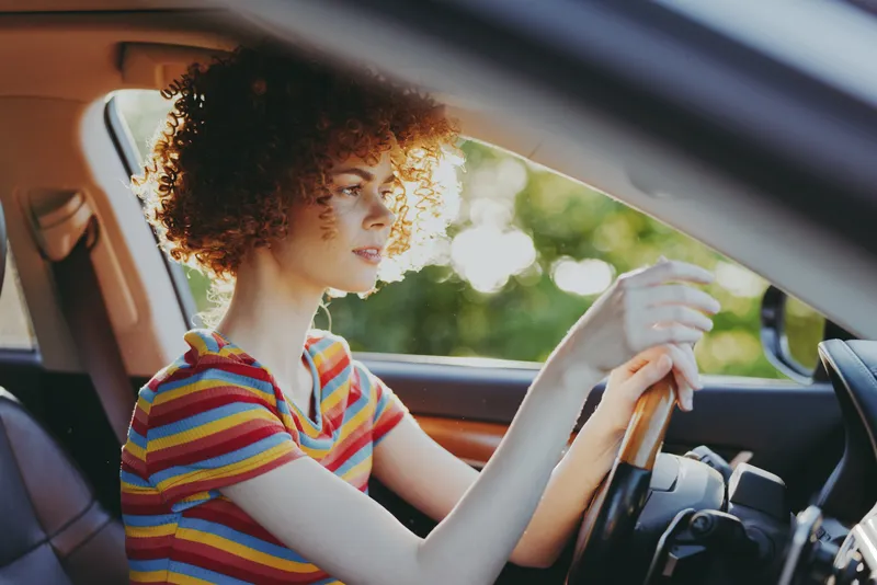 Young woman with curly hair drives a car in a sunlit moment, wearing a striped shirt and focused on the road.