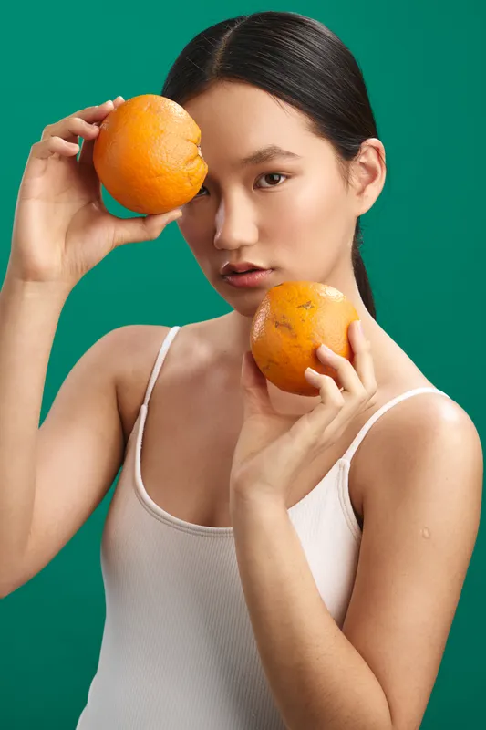 Asian woman holds two bright oranges against a vivid green backdrop, creating a fresh still life. Her calm expression and direct gaze convey health, energy, and a modern studio vibe.
