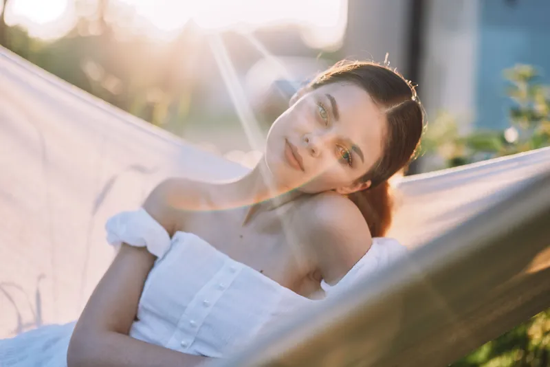 Young woman relaxing in a hammock during sunset, wearing a white dress, embodying tranquility and natural beauty, with soft sunlight and nature background