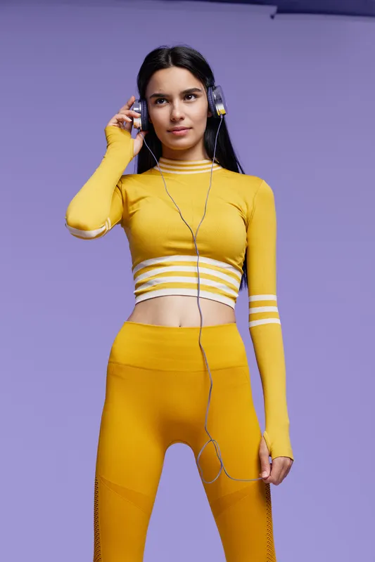 sport fitness woman in yellow activewear with headphones listens to music, focused and poised, standing against purple background, healthy lifestyle and workout concept