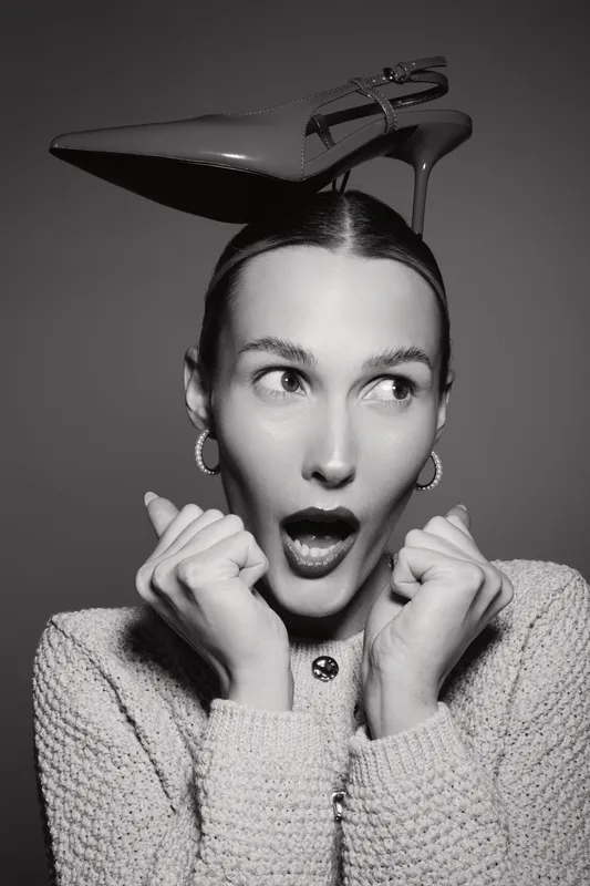 Surprised white woman with fair skin wears a high heel shoe on her head, looking sideways with open mouth and hands near face, wearing textured sweater and hoop earrings.