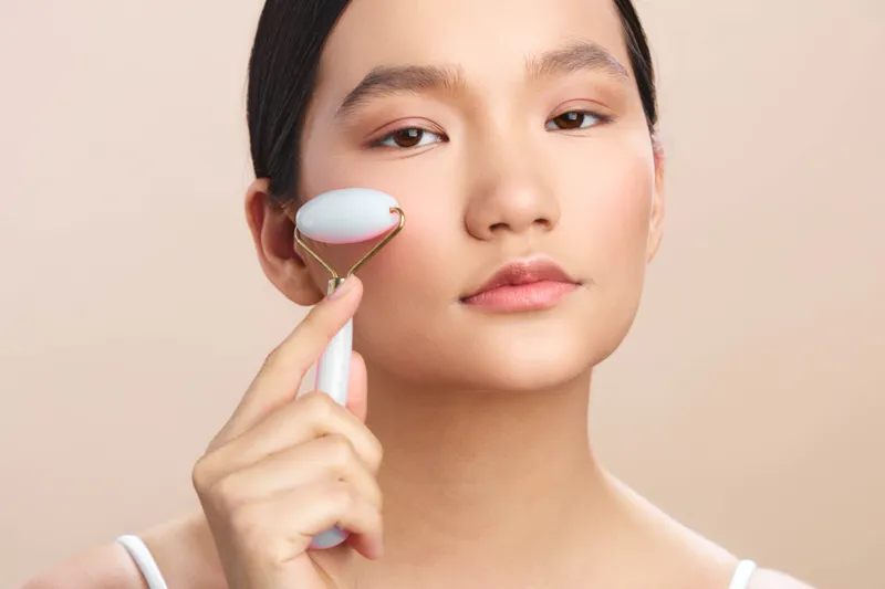 Asian woman gently uses a facial roller in a serene skincare routine, with soft studio lighting and a calm, focused expression creating an intimate wellness moment.