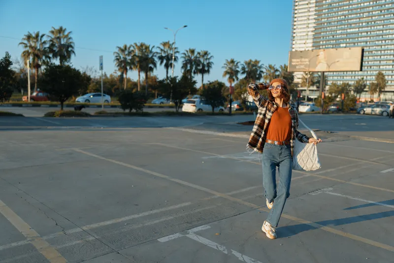 Woman walking in urban parking lot wearing sunglasses and casual outfit, carrying tote bag in warm sunlight and palm-lined city scene, candid lifestyle portrait with golden hour glow and authenticity.