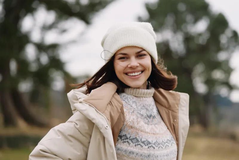 Happy woman outdoors wearing a beige winter coat, knit sweater and white beanie, enjoying a breezy day in a park with soft blurred trees and open sky.
