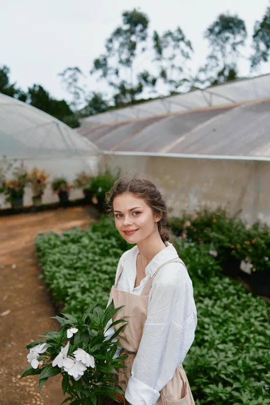 Young female florist in a greenhouse, wearing a light colored apron, holding fresh flowers, showcasing a serene smile amidst lush greenery