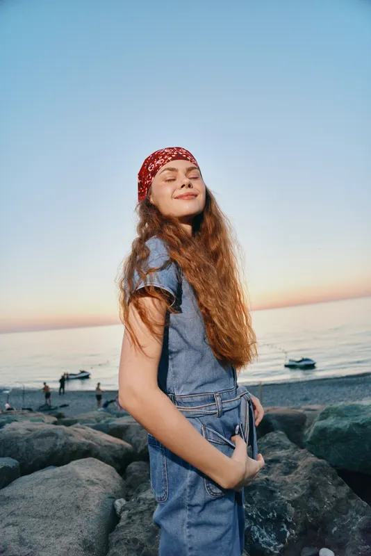 Lifestyle woman in modern boho-western denim street style with relaxed smile by the seaside during golden hour, film color capturing natural light and casual attitude.